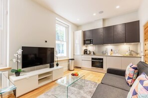 A modern living room with a flat-screen TV, a glass coffee table, a sofa, and a kitchen area with dark cabinets and a microwave. - Super Central Lovely 1BR Flat - Fitzrovia (London)