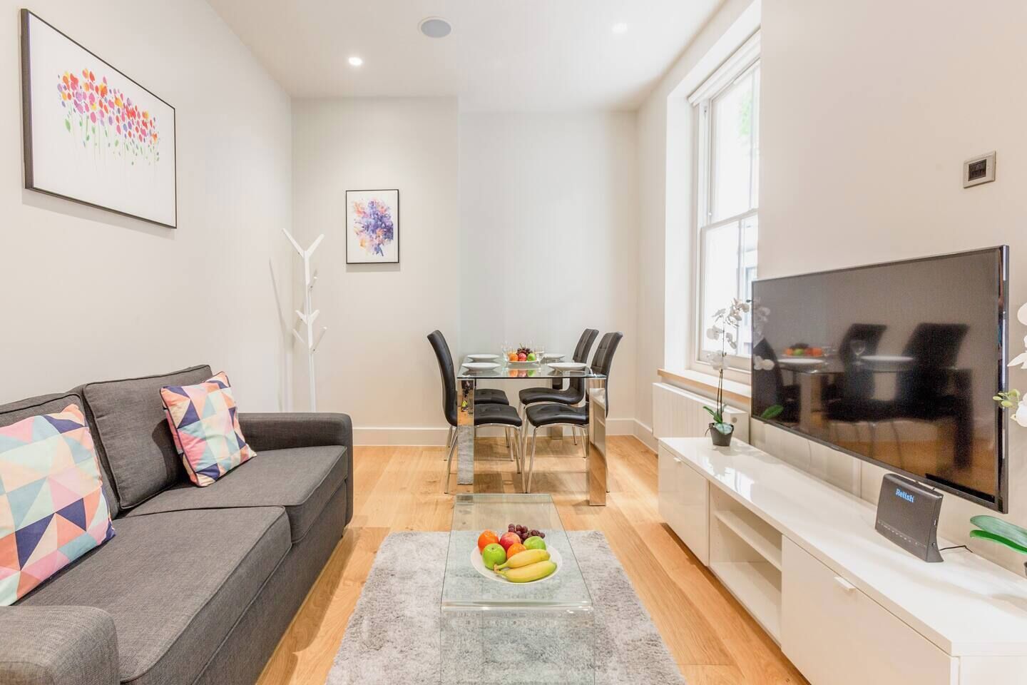 A modern living room with a grey sofa, a dining table with chairs, a flat-screen TV, and artwork on the walls.