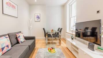 A modern living room with a grey sofa, a dining table with chairs, a flat-screen TV, and artwork on the walls.