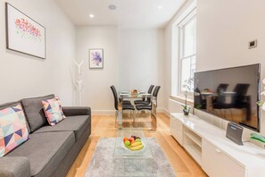A modern living room with a grey sofa, a dining table with chairs, a flat-screen TV, and artwork on the walls. - BnBNova - 1BR - Soho & Central London (London)