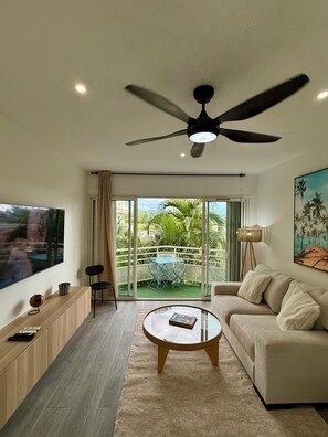 A modern living room with a ceiling fan, a large TV, a sofa, a coffee table, and a view of a balcony with greenery. - cosy apartment (saint martin)