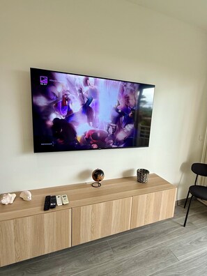 A flat-screen TV mounted on a wall displaying a music video. Below the TV, there's a wooden console with a globe, remote controls, and decorative items. - cosy apartment (saint martin)