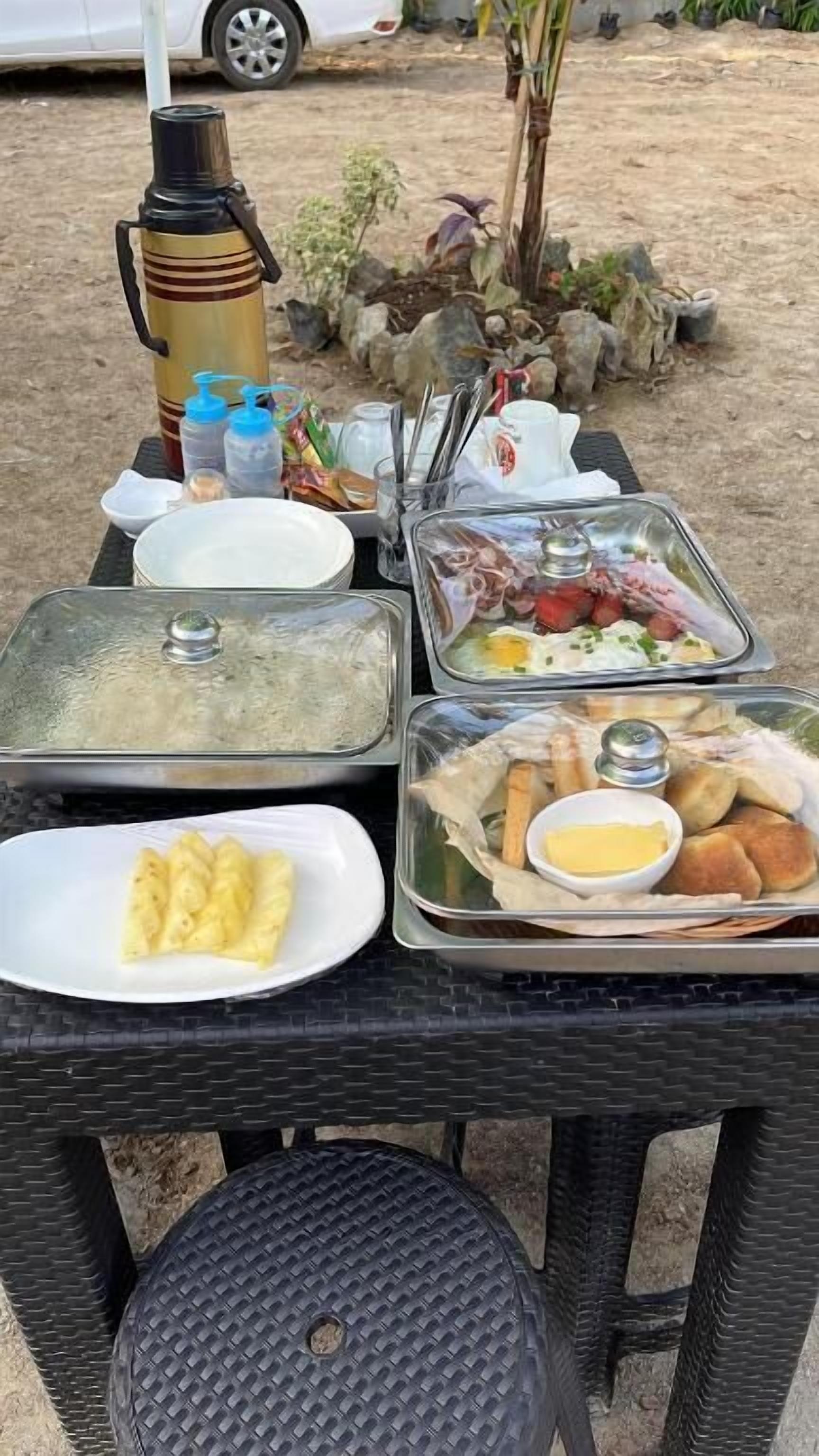 Una mesa con diversos alimentos y un termo, ubicada al aire libre junto a una silla y otra mesa pequeña.