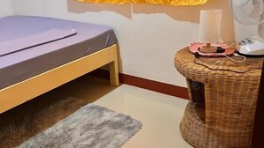 Una habitación pequeña con una cama, un ventilador, una silla de mimbre y una alfombra.