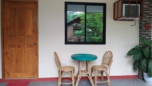 Una habitación con puerta de madera, ventana con vista a follaje, una pequeña mesa redonda con sillas y una planta en maceta.