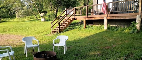 Una terraza de madera con una escalera que lleva a una zona cubierta, un hoyo para la fogata y sillas blancas de plástico.