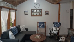 A cozy living room with a wooden ceiling, a sofa with a 'Farm to Table' pillow, two chairs with 'Bovina' blankets, a coffee table with a magazine, and a heart-shaped light fixture on the wall.