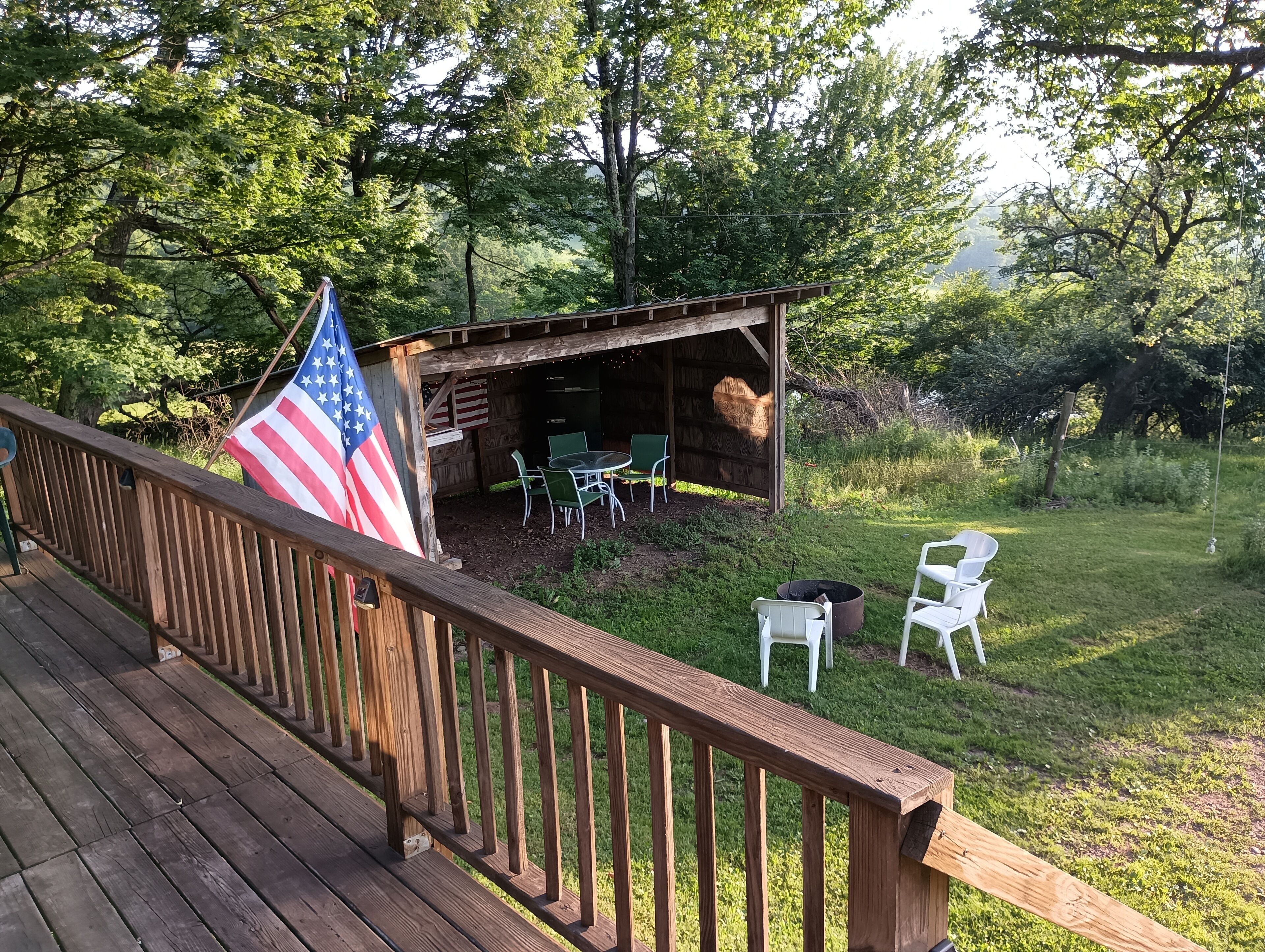 A wooden deck with a railing, an American flag, a covered outdoor seating area with tables and chairs, and a grassy yard.