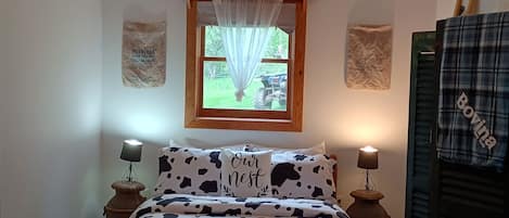 Un dormitorio con una cama de madera, dos lámparas de noche, un ventanal con cortinas traslúcidas y una repisa con botellas.