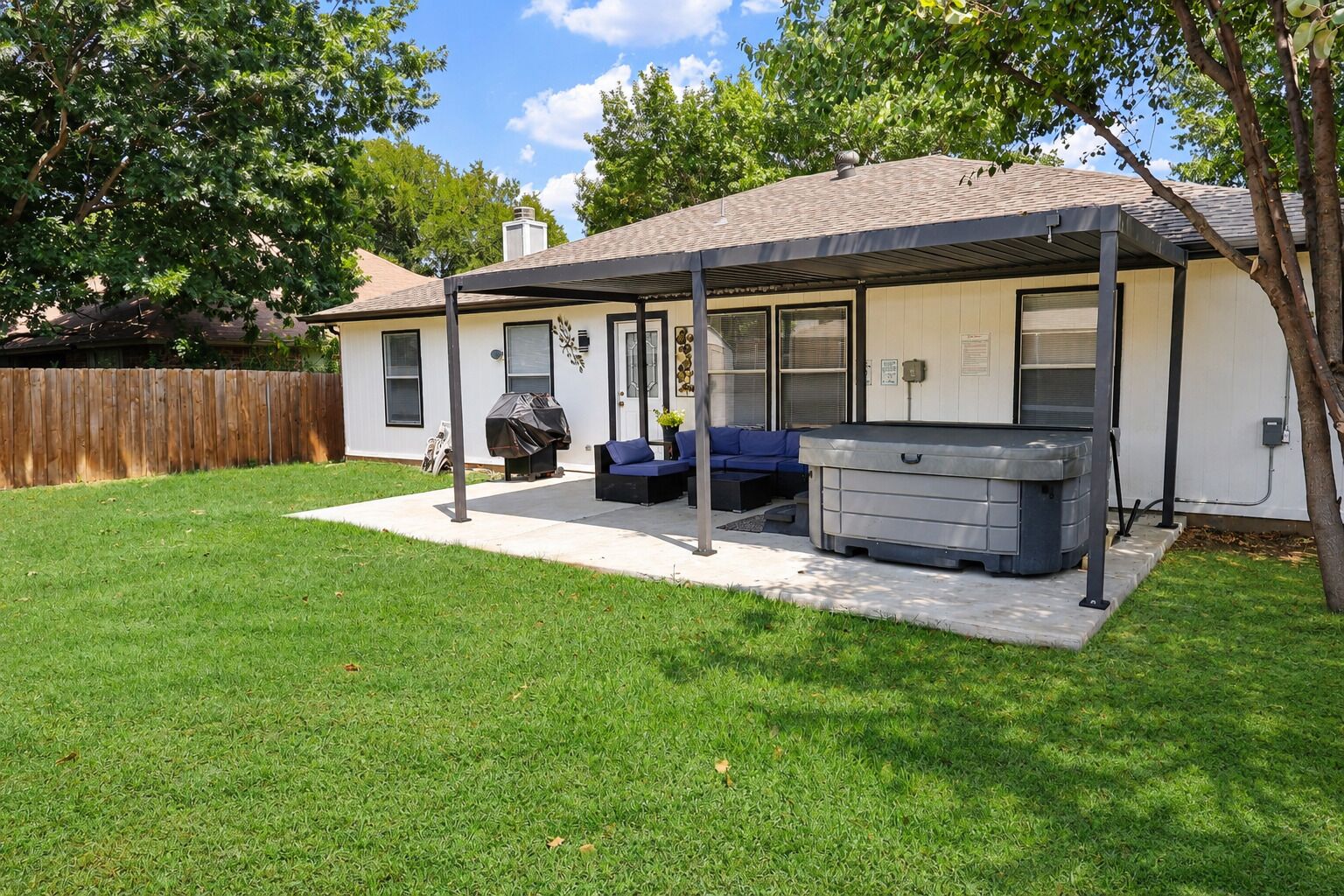 A covered patio area with a hot tub, outdoor seating, and a wooden fence in the background.