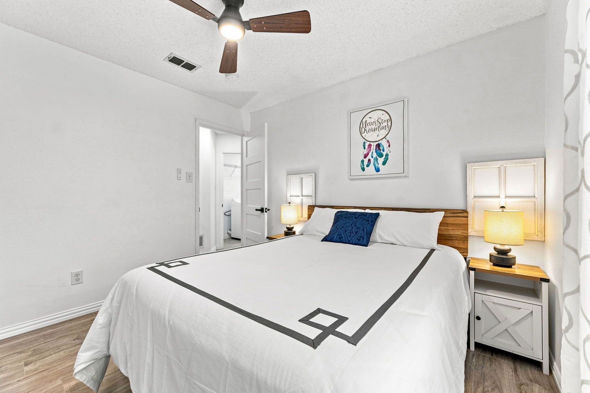 Une chambre à coucher comprenant un lit, une table de chevet, une lampe, un ventilateur de plafond et un mur orné d’un attrape-rêves.