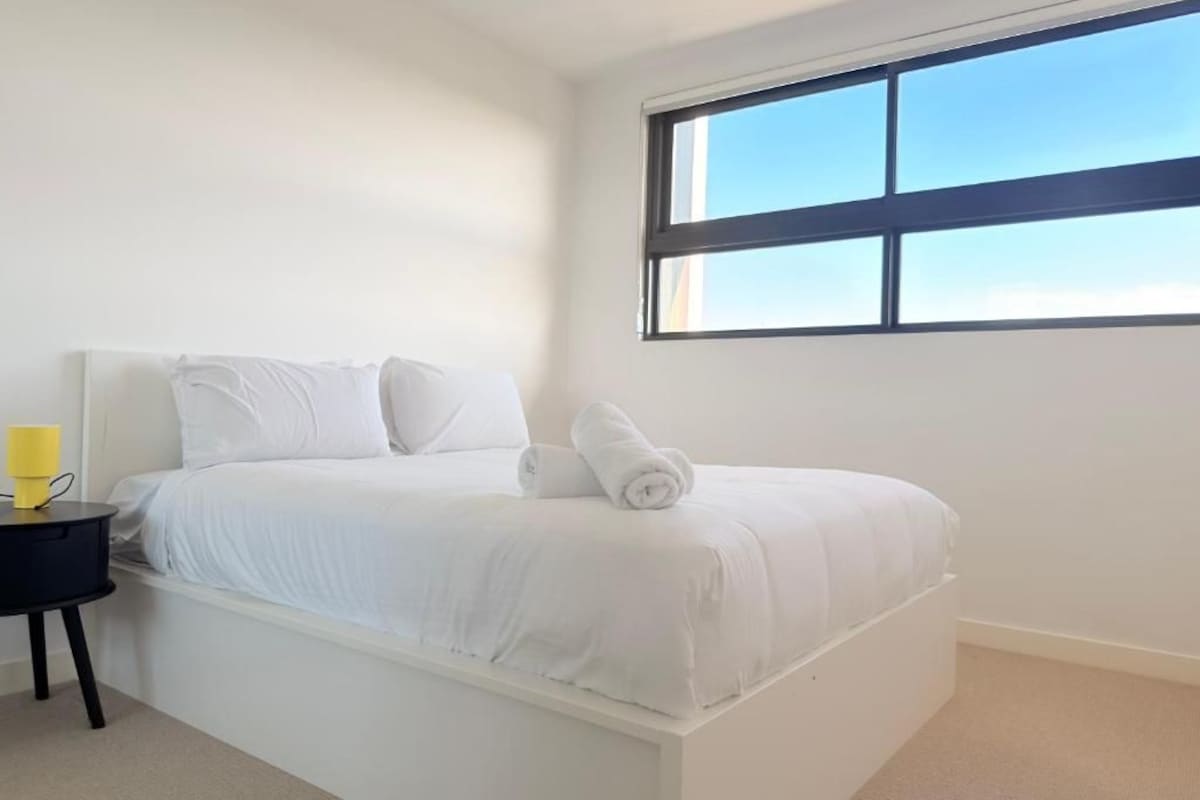 A white bed with a rolled towel, a black nightstand with a yellow object, and a window with a view of the sky.