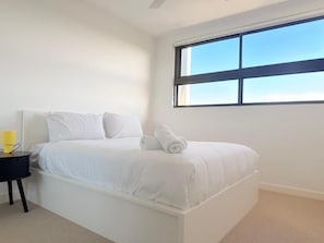 A white bed with a rolled towel, a black nightstand with a yellow object, and a window with a view of the sky.