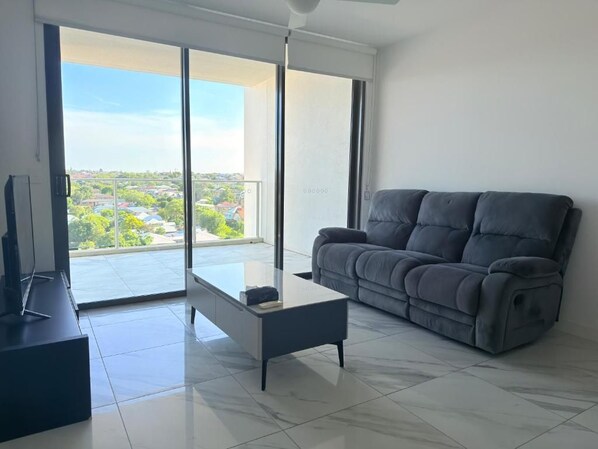 A modern living room with a grey sofa, a glass coffee table, and a view through large sliding glass doors.