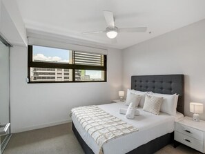 A modern bedroom with a bed, bedside tables, a ceiling fan, and a window with blinds. - Timi Property (Woolloongabba)