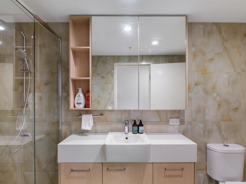 A bathroom with a glass-enclosed shower, a white sink with two faucets, a mirror above the sink, and a toilet to the right.