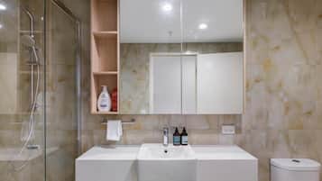 Une salle de bain avec une douche à parois de verre, un lavabo blanc doté de deux robinets, un miroir au-dessus du lavabo et des toilettes situées à droite.
