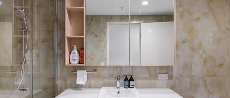 A bathroom with a glass-enclosed shower, a white sink with two faucets, a mirror above the sink, and a toilet to the right.