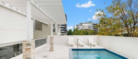 A rooftop swimming pool with a clear blue pool, white lounge chairs, and a view of surrounding buildings.