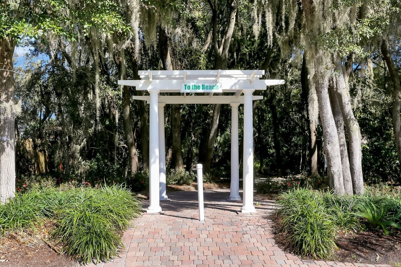 Eine weiße Pergola mit einem Schild, auf dem 'Zum Strand' steht, in einem Garten mit Bäumen und Pflanzen.