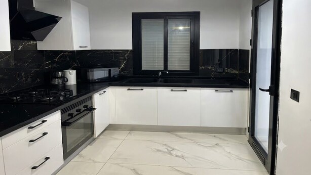 A modern kitchen with a black countertop, white cabinets, a built-in oven, and a window with blinds.