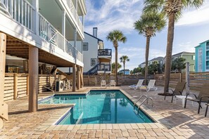 A swimming pool with a wooden deck and lounge chairs, surrounded by palm trees and a multi-story building with balconies. - Barefoot Dreams: Ocean View & Pool (Surfside Beach)