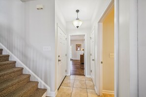 A hallway with a carpeted staircase, white walls, a tiled floor, and a hanging light fixture. - Crenshaw Corner: Coastal Charm w/ Shared Pool, Steps 2 Beach (Murrells Inlet)