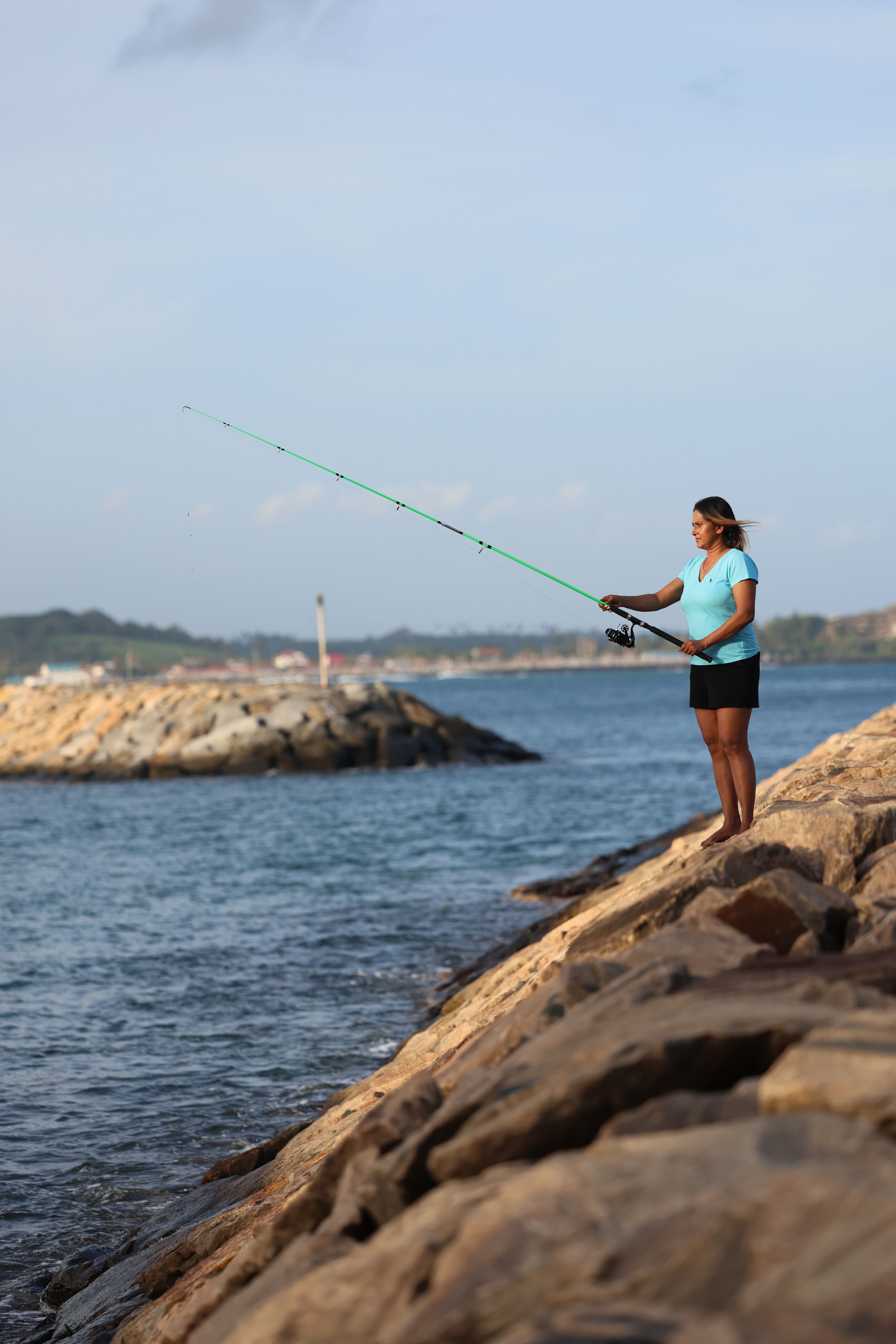 A person fishing on a rocky shore with a body of water and a distant lighthouse.