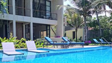 A swimming pool with lounge chairs, a building with balconies, and tropical vegetation.