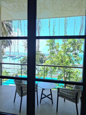 Habitación con vistas a una playa y al océano, que incluye un ventanal grande, dos sillas de mimbre y una mesita.