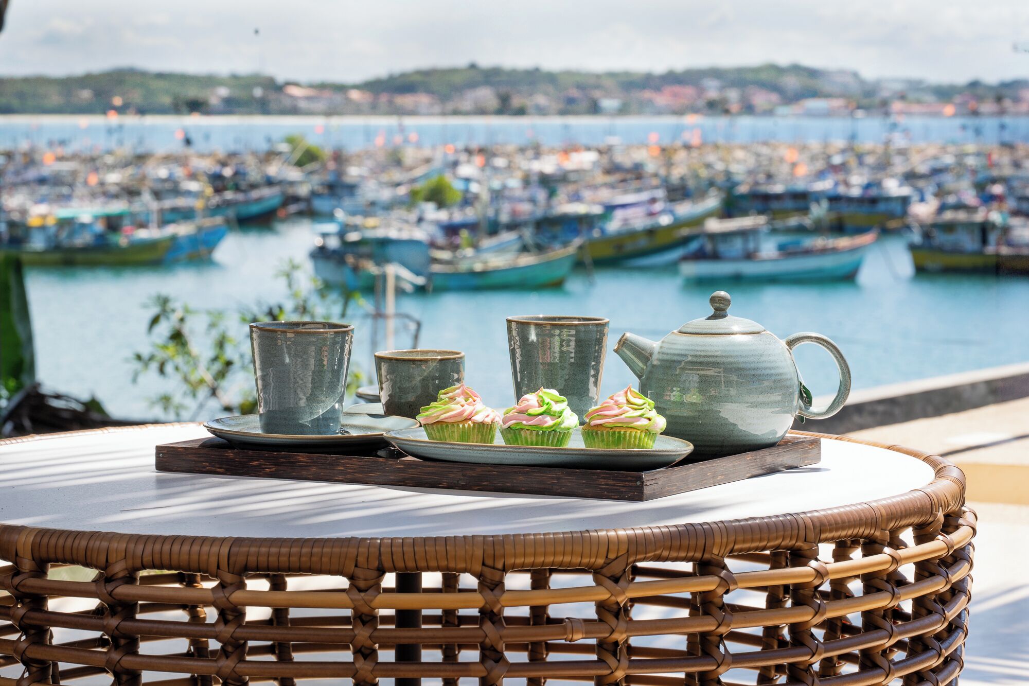 Une table en osier, sur laquelle sont disposés une théière, des tasses et des petits gâteaux, se trouve sur une terrasse donnant sur un port où sont amarrés des bateaux.
