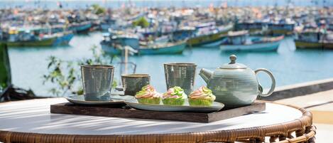 Une table en osier, sur laquelle sont disposés une théière, des tasses et des petits gâteaux, se trouve sur une terrasse donnant sur un port où sont amarrés des bateaux.