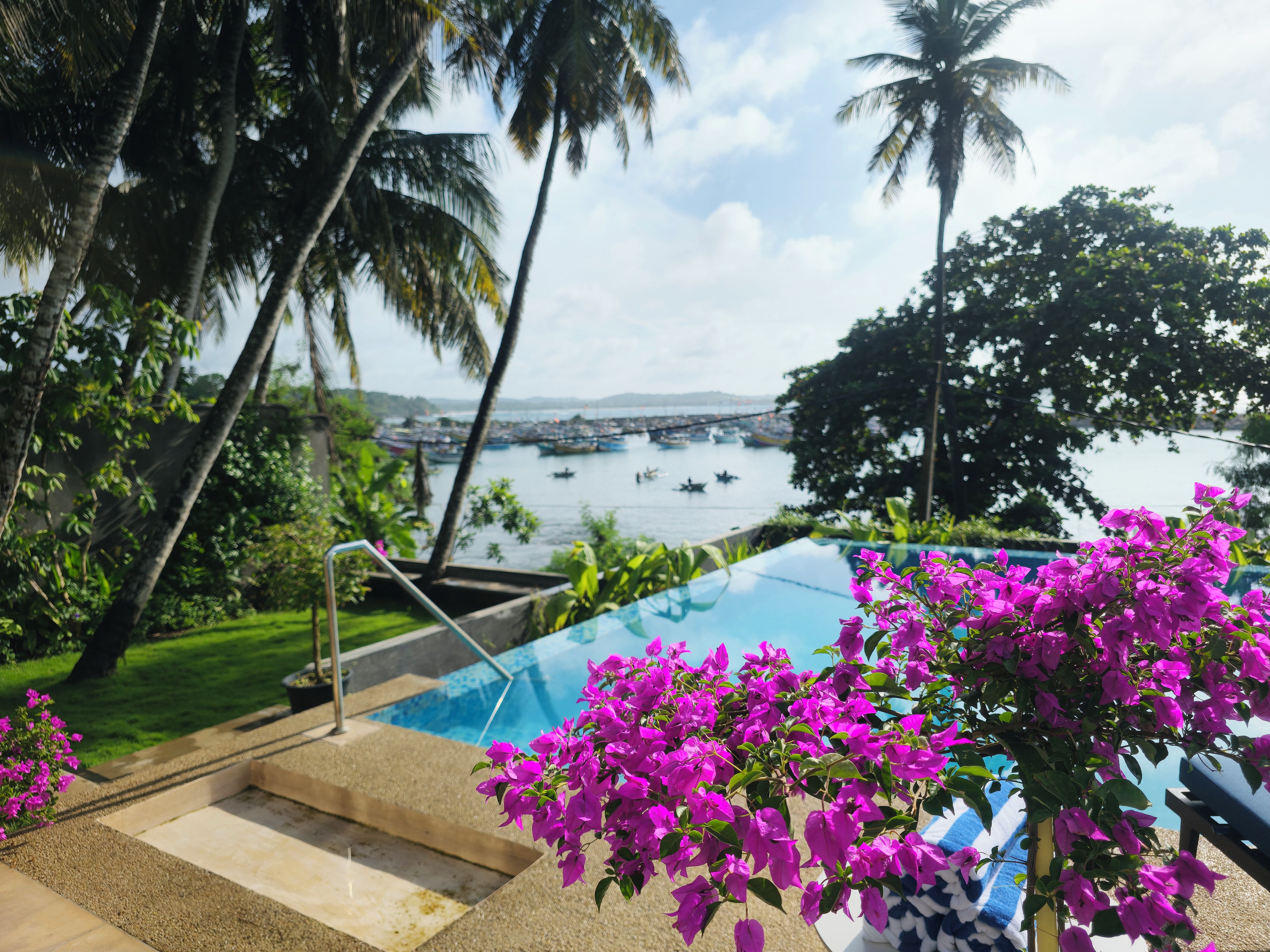 A pool area with a view of boats on water, surrounded by palm trees and greenery.
