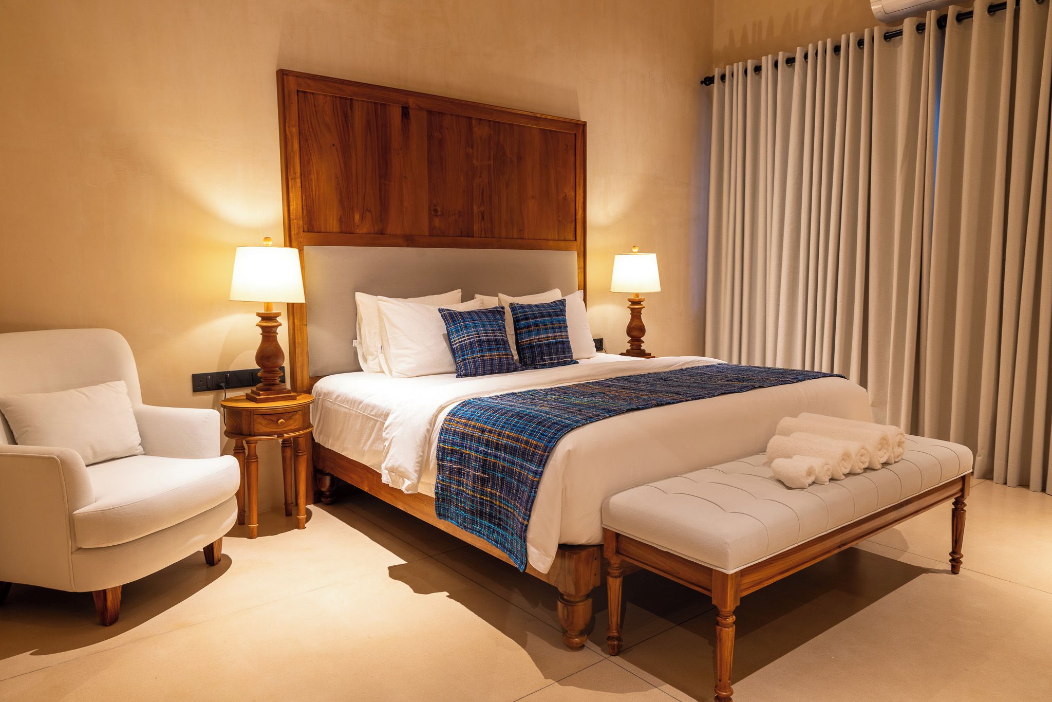 A hotel room with a large bed, two bedside tables with lamps, a chair, and a bench.