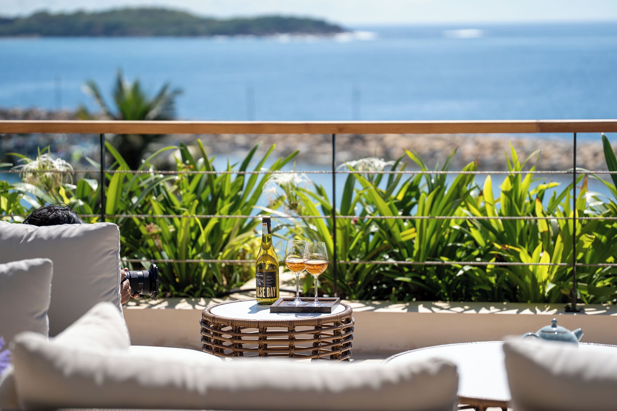 A balcony with a view of the sea, a wicker table with a bottle and glasses, and white cushioned chairs.