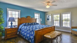 A bedroom with a wooden bed, a ceiling fan, and a view through large windows. - Crenson Beach: Steps from the Beach & Pool (Murrells Inlet)