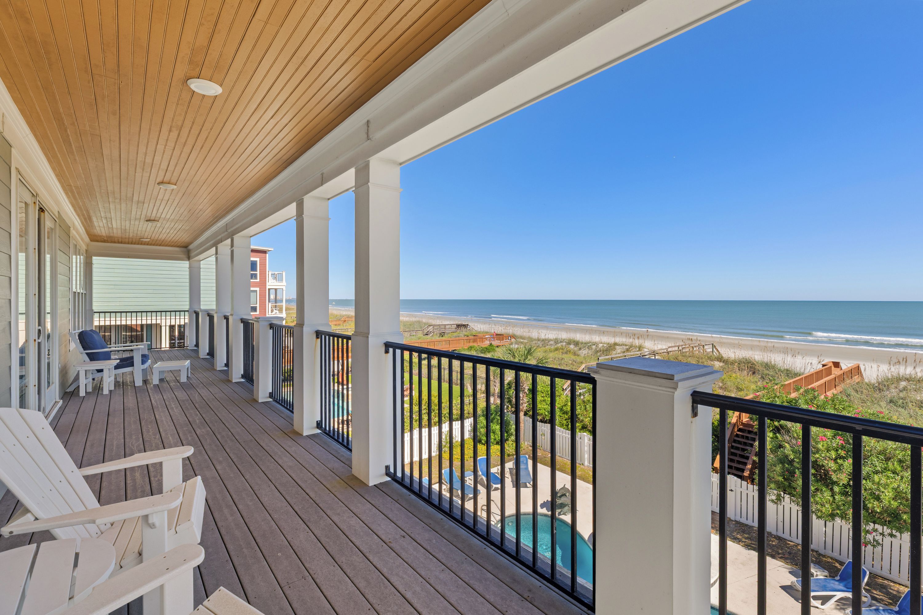 A wooden deck with white chairs, a pool, and a view of the beach.