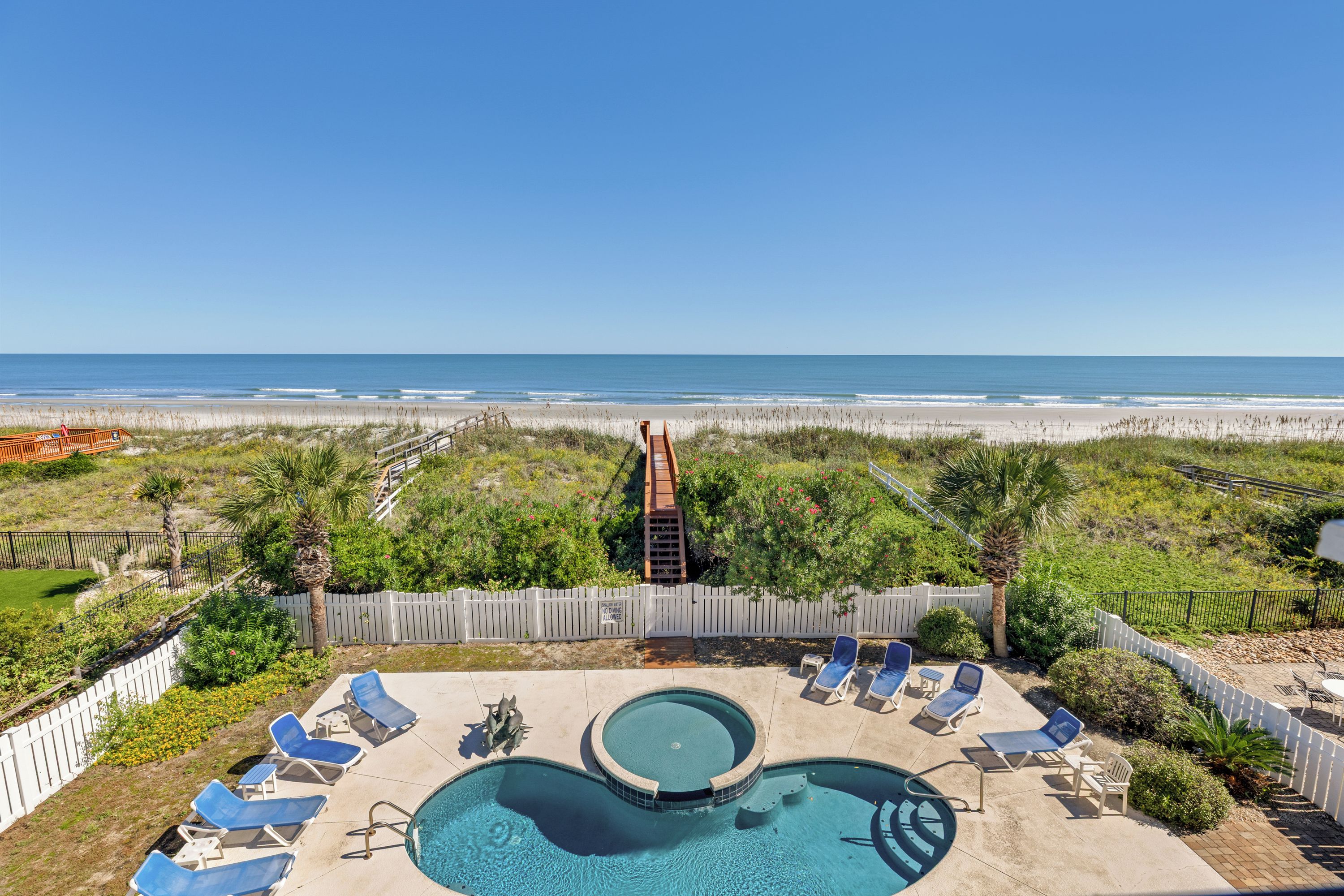 A pool area with a view of the beach, surrounded by a fence and palm trees.