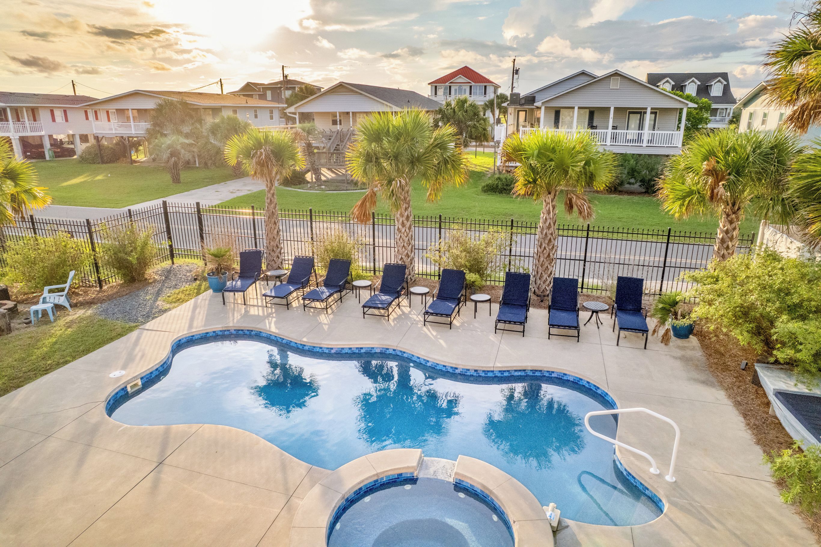 A swimming pool with lounge chairs and a small hot tub, surrounded by a fence and palm trees.