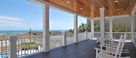A covered outdoor deck with white railings and wooden flooring, overlooking a beach and ocean.