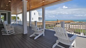 A covered outdoor deck with white rocking chairs, a view of the ocean, and a white building in the background. - Gulf Stream Breeze: Oceanfront Luxury, Private Pool & Outdoor Oasis (Murrells Inlet)
