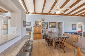 A spacious kitchen with a dining area, wooden ceiling beams, and various framed artworks on the walls. - Panacea: Oceanfront Charm with Pool, & Private Beach Walkway (Murrells Inlet)