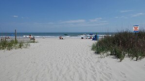 A sandy beach with a clear sky, a sign indicating to keep off dunes, and a few people and umbrellas in the distance. - Quality Time: Private Pool Retreat Just Steps from the Beach (Murrells Inlet)