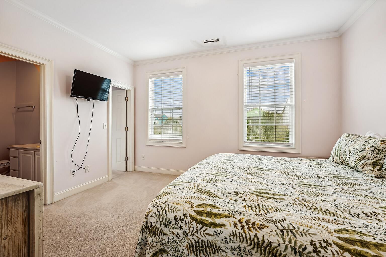 A bedroom with a bed, a TV mounted on the wall, and two windows with blinds.