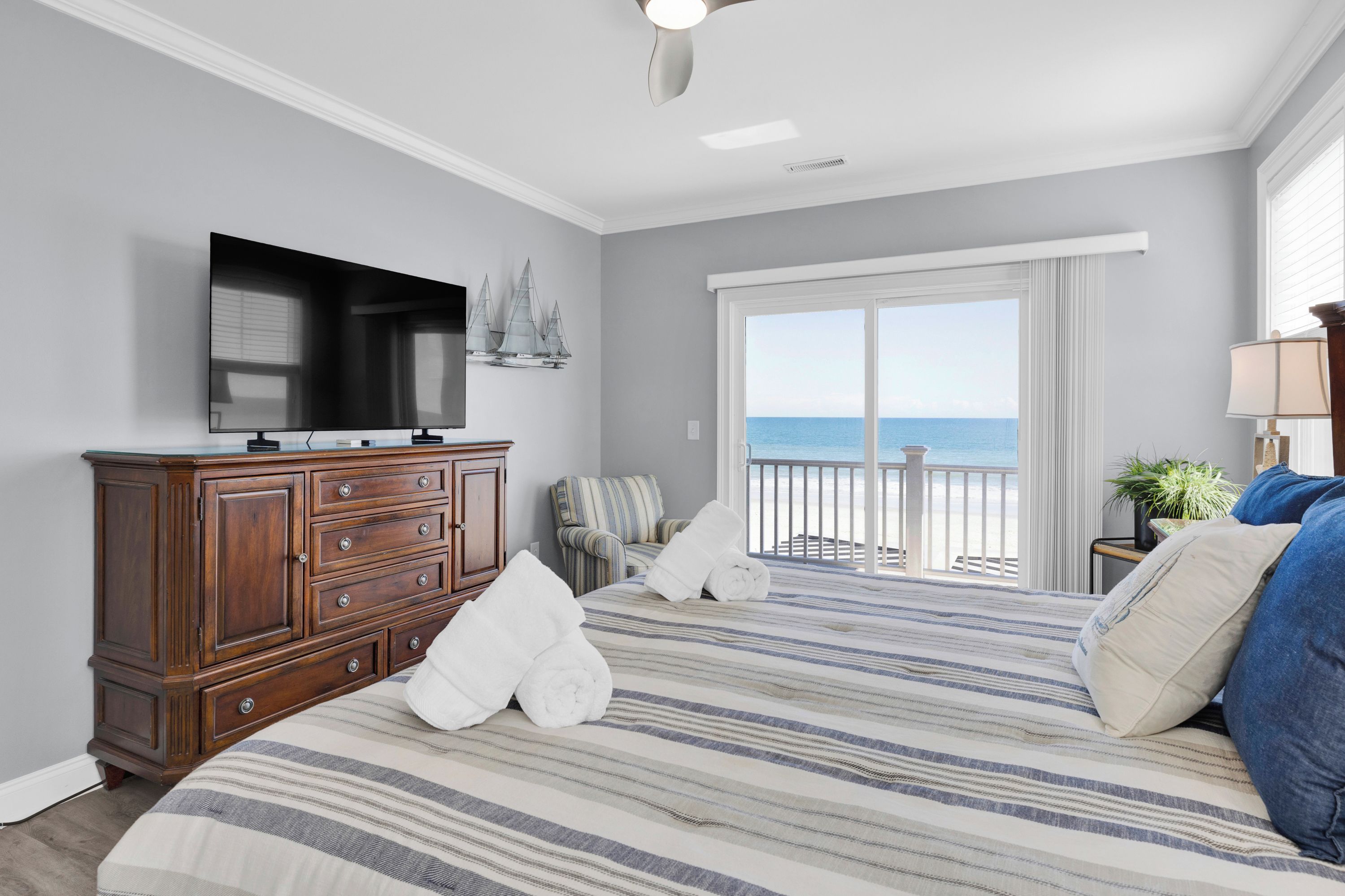 A bedroom with a bed, a dresser, a television, a chair, and a view of the ocean through a large window.