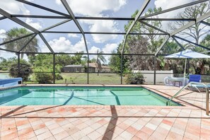A swimming pool with a tiled deck, surrounded by a metal fence, and a view of a residential area with trees and a house in the background. - Rotonda West Getaway | Private Heated Pool Retreat (Rotonda West)