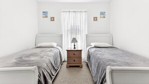 Un dormitorio con dos camas individuales, una mesita de noche, una lámpara y una ventana con cortinas translúcidas.