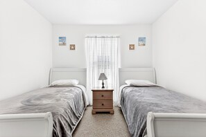 Ein Schlafzimmer mit zwei Einzelbetten, einem Nachttisch, einer Lampe und einem Fenster mit leichtem Vorhang.