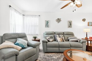 Een woonkamer met een grijze bank, een leren fauteuil, een glazen salontafel en een plafondventilator.