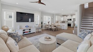A modern living room with a flat-screen TV, a dining area with a table and chairs, and a kitchen with appliances. - Upscale New Construction 4BR/3BA Coastal Retreat • 3-Min Walk to Beach •Sleeps 9 (Carolina Beach)